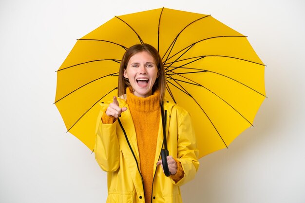 Foto jovem inglesa com casaco à prova de chuva e guarda-chuva isolado no fundo branco surpreso e apontando para a frente