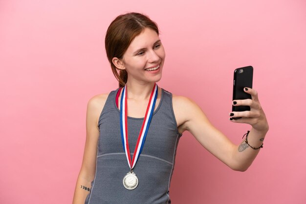 Jovem inglesa com medalhas isoladas em fundo rosa fazendo uma selfie