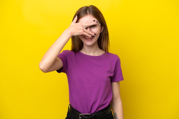 Jovem inglesa isolada em fundo amarelo cobrindo os olhos com as mãos e sorrindo