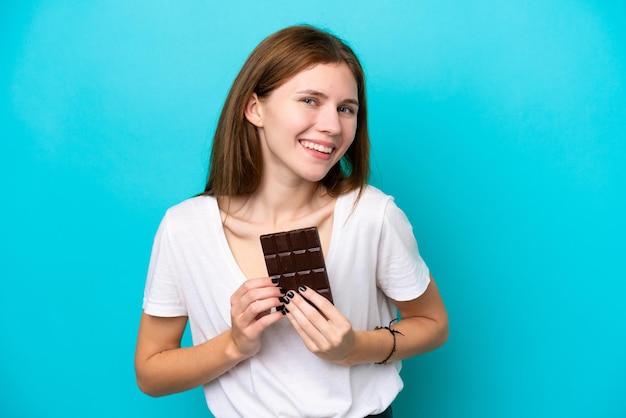 Jovem inglesa isolada em fundo azul tomando uma tablete de chocolate e feliz