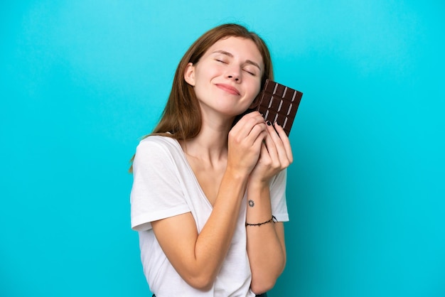 Foto jovem inglesa isolada em fundo azul tomando uma tablete de chocolate e feliz