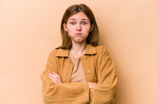 Jovem inglesa isolada em fundo bege sopra bochechas, tem expressão cansada. Conceito de expressão facial.