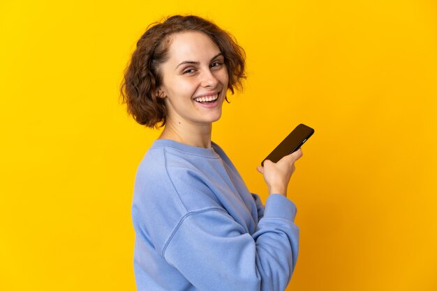 Jovem inglesa isolada na parede amarela segurando um telefone celular e com os braços cruzados