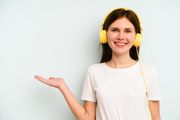 Jovem inglesa ouvindo música isolada em fundo azul, mostrando um espaço de cópia na palma da mão e segurando outra mão na cintura