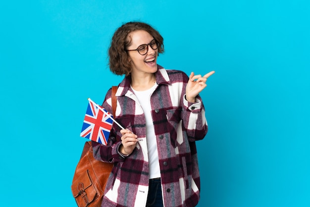 Jovem inglesa segurando uma bandeira do Reino Unido isolada em um fundo azul, apontando o dedo para o lado e apresentando um produto