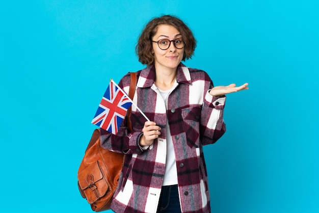 Jovem inglesa segurando uma bandeira do Reino Unido, isolada em uma parede azul, tendo dúvidas ao levantar as mãos