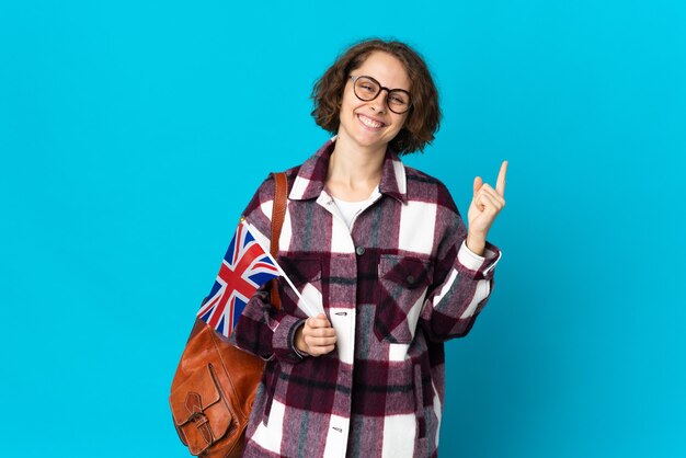 Jovem inglesa segurando uma bandeira do Reino Unido isolada no espaço azul, mostrando e levantando um dedo em sinal dos melhores