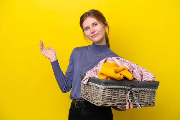 Jovem inglesa segurando uma cesta de roupas isolada em fundo amarelo, estendendo as mãos para o lado para convidar para vir