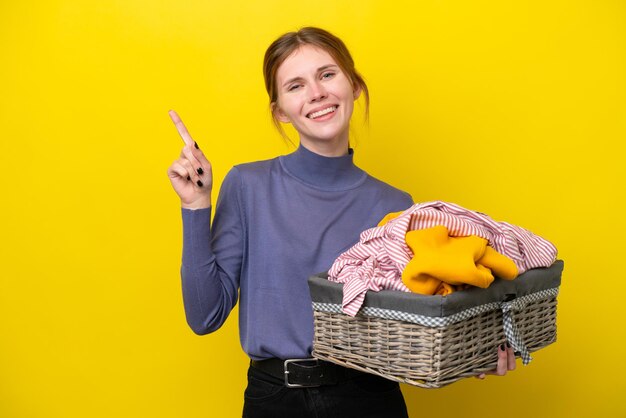 Jovem inglesa segurando uma cesta de roupas isolada em fundo amarelo mostrando e levantando um dedo em sinal do melhor