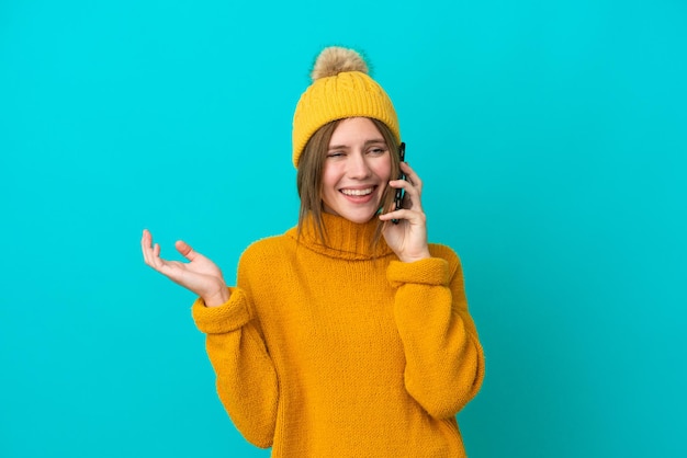 Jovem inglesa vestindo jaqueta de inverno isolada em fundo azul, mantendo uma conversa com o celular com alguém