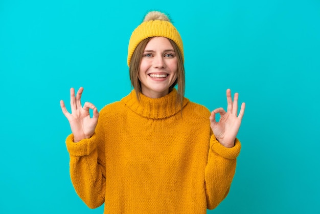 Jovem inglesa vestindo jaqueta de inverno isolada em fundo azul, mostrando um sinal de ok com os dedos