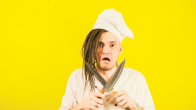 Jovem insano vestido de chef com facas em fundo amarelo Cozinheiro maluco de chapéu branco e camisa posando com facas de cozinha