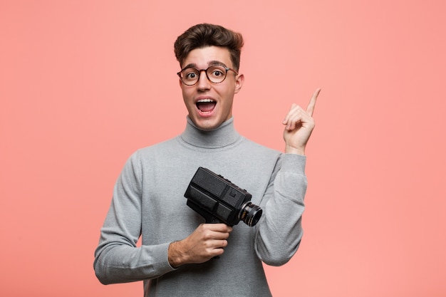 Jovem intelectual segurando uma câmera de filme sorrindo alegremente apontando com o dedo indicador.