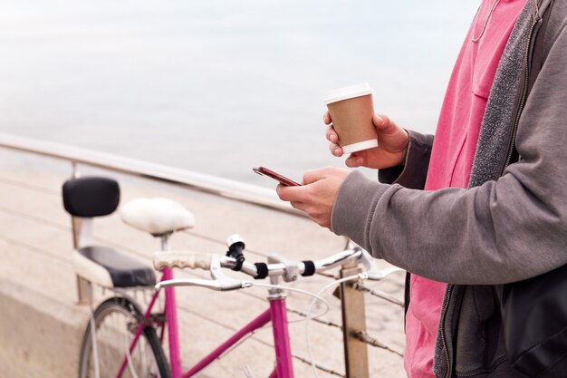 Jovem irreconhecível com uma bebida, usando um telefone celular ao lado de uma bicicleta vintage