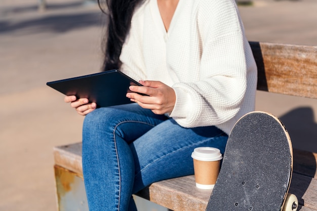 Jovem irreconhecível sentada em um banco usando seu tablet ao lado de um café e um skate