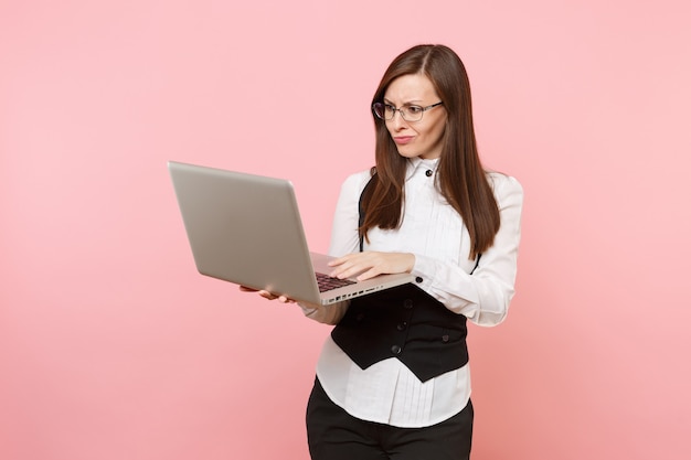 Jovem irritada mulher de negócios insatisfeita em copos, trabalhando no computador laptop pc isolado em fundo rosa pastel. Senhora chefe. Conceito de riqueza de carreira de conquista. Copie o espaço para anúncio.