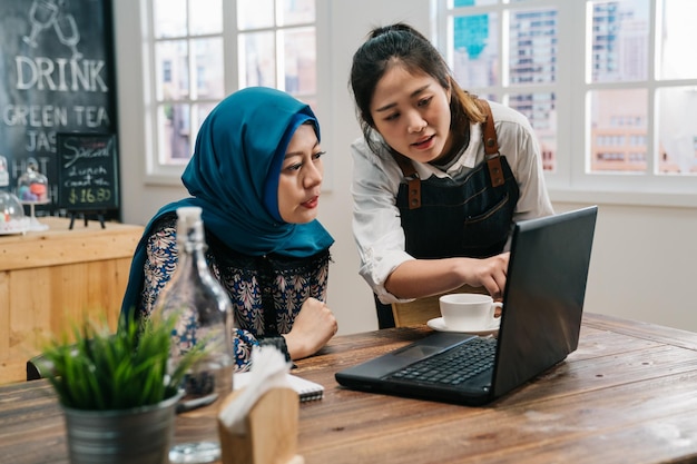 jovem islão freelancer feminino usando computador portátil enquanto aguarda o pedido. garçonete de garota amigável no avental servindo café para convidados no café bar moderno. duas mulheres cliente e amigos da equipe discutindo