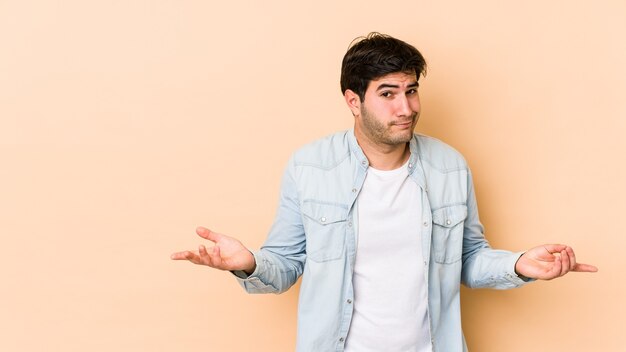 Jovem isolado em um fundo bege duvidando e encolhendo os ombros em gesto de questionamento.