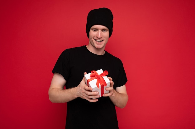jovem isolado sobre a parede de fundo vermelho, usando chapéu preto e camiseta preta, segurando uma caixa de presente branca com fita vermelha e olhando para a câmera.