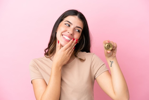 Jovem italiana segurando um bitcoin isolado em fundo rosa olhando para cima enquanto sorria