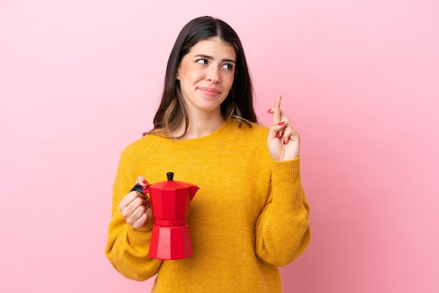 Jovem italiana segurando uma cafeteira isolada em fundo rosa com dedos cruzando e desejando o melhor