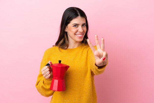 Jovem italiana segurando uma cafeteira isolada em fundo rosa feliz e contando três com os dedos