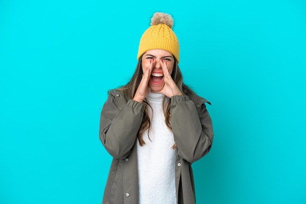 Jovem italiana vestindo jaqueta de inverno e chapéu isolado em fundo azul gritando e anunciando algo