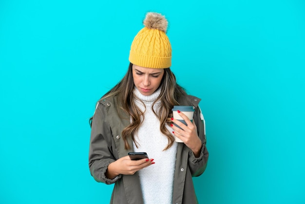 Jovem italiana vestindo jaqueta de inverno e chapéu isolado em fundo azul segurando café para levar e um celular