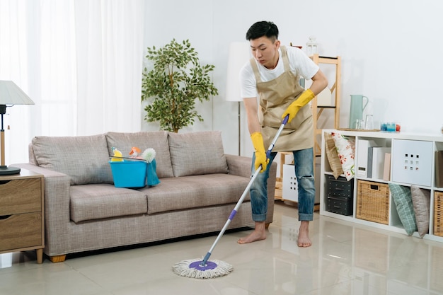 Jovem japonês asiático vestindo avental de limpeza de chão em casa. cara lavando o chão com esfregão e balde na sala de estar do apartamento moderno brilhante. família masculina ajudando a esposa a arrumar a casa.