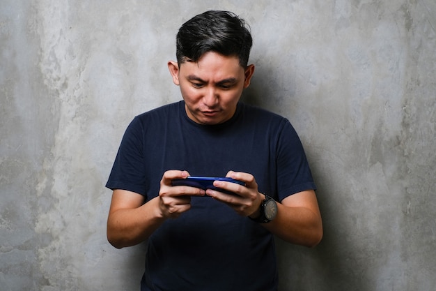 Jovem japonês usando smartphone em pé sobre o fundo da parede de concreto usando o smartphone jogando jogo para celular sobre o fundo da parede de concreto