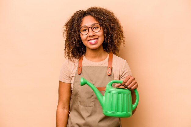 Jovem jardineira afro-americana segurando irrigação isolada em fundo bege feliz sorrindo e alegre