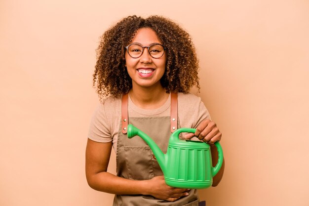 Jovem jardineira afro-americana segurando irrigação isolada em fundo bege rindo e se divertindo