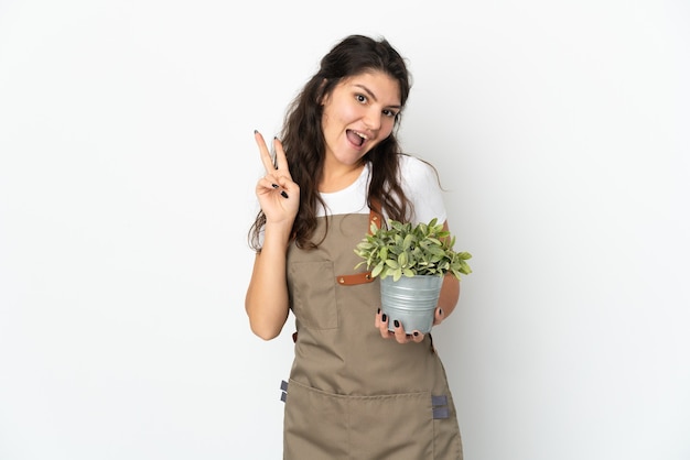 Jovem jardineira russa segurando uma planta isolada, sorrindo e mostrando o sinal da vitória