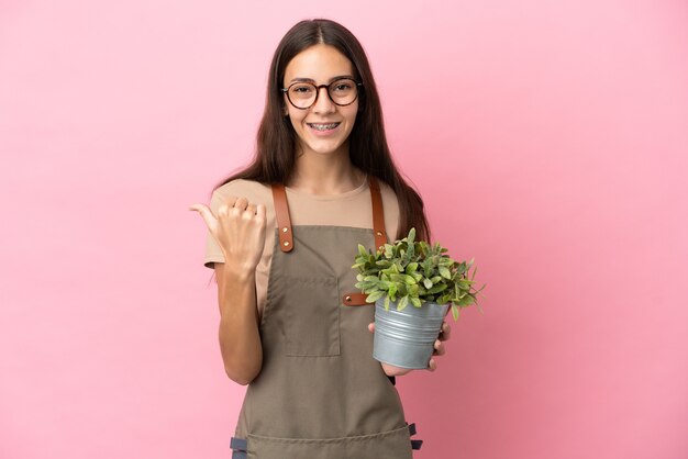 Jovem jardineira segurando uma planta isolada em um fundo rosa apontando para o lado para apresentar um produto