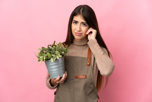 Jovem jardineira segurando uma planta isolada em um fundo rosa frustrada e cobrindo as orelhas