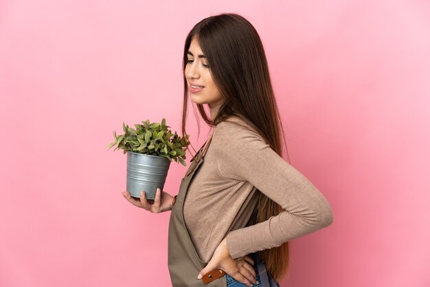 Jovem jardineira segurando uma planta isolada na parede rosa, sofrendo de dor nas costas por ter feito um esforço