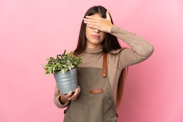 Jovem jardineira segurando uma planta isolada no fundo rosa, cobrindo os olhos com as mãos. Não quero ver nada