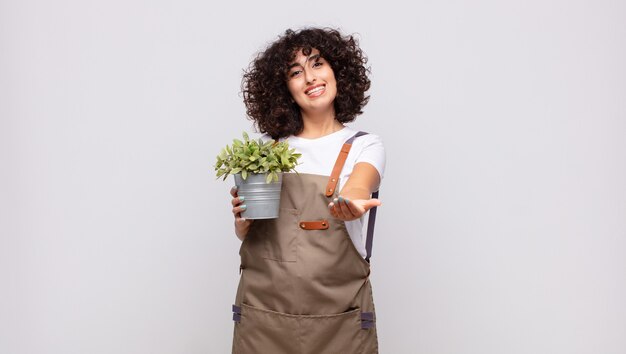 Jovem jardineira sorrindo feliz com um olhar amigável, confiante e positivo, oferecendo e mostrando um objeto ou conceito