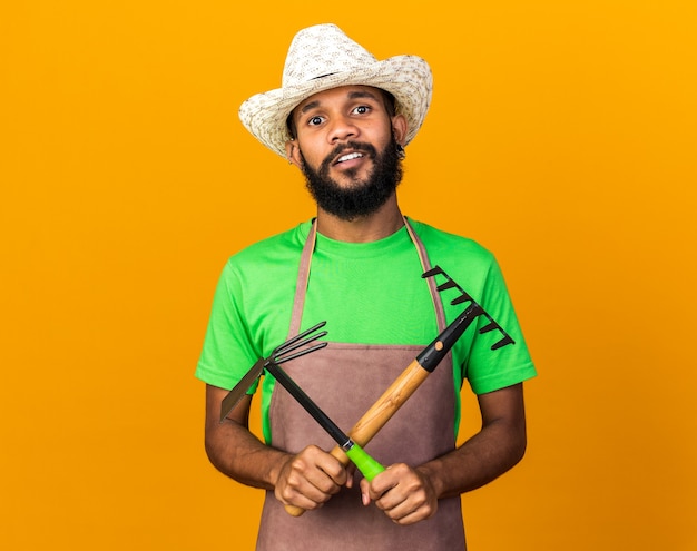 Jovem jardineiro afro-americano satisfeito com um chapéu de jardinagem, segurando e cruzando o ancinho com o ancinho de enxada