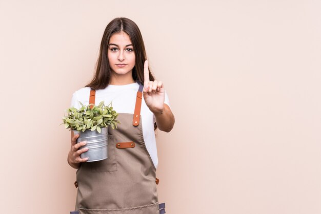 Jovem jardineiro caucasiana mulher segurando uma planta isolada; mostrando o número um com o dedo.