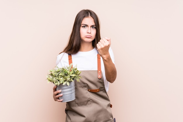 Jovem jardineiro mulher caucasiana, segurando uma planta isolada, mostrando o punho, expressão facial agressiva.