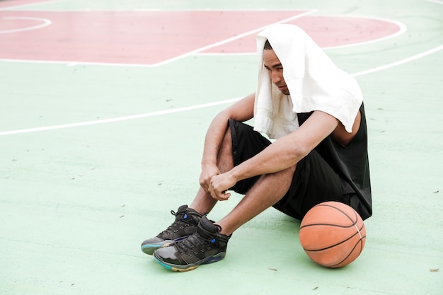 Jovem jogador de basquete feliz sentado no parque com uma toalha