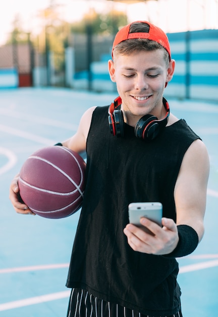 Jovem jogador de basquete com uma bola olha para seu celular e sorri enquanto faz uma pausa