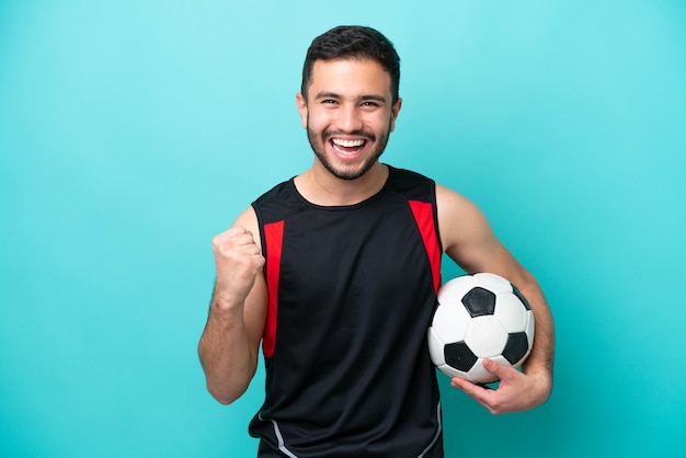 Jovem jogador de futebol brasileiro isolado em fundo azul comemorando uma vitória na posição de vencedor