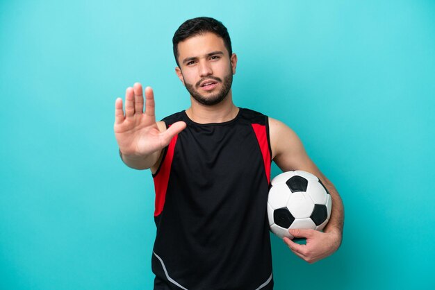 Jovem jogador de futebol brasileiro isolado em fundo azul fazendo gesto de parada