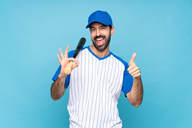 Jovem jogando beisebol sobre azul isolado mostrando sinal bem e polegar para cima gesto