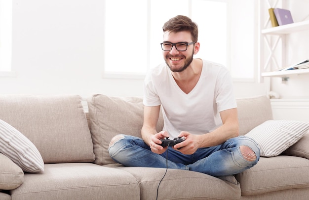 Jovem jogando videogame em casa. Um cara feliz de óculos sentado no sofá com o joystick nas mãos, copie o espaço