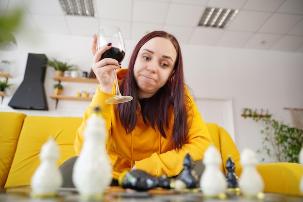 Jovem jogando xadrez e bebendo vinho tinto no quarto Mulher adulta com álcool joga no jogo de tabuleiro lógico consigo mesma sentada no sofá