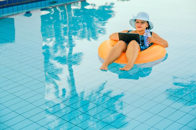 Jovem judia de chapéu de sol na piscina no círculo de natação com um coquetel e um laptop