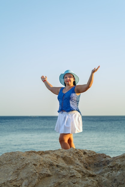 Jovem judia de saia branca e chapéu azul à beira-mar perto das rochas feliz Vertical